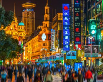 Busy neon-lit city street at night with tall colorful signs and blurred pedestrians getting by the storefronts.