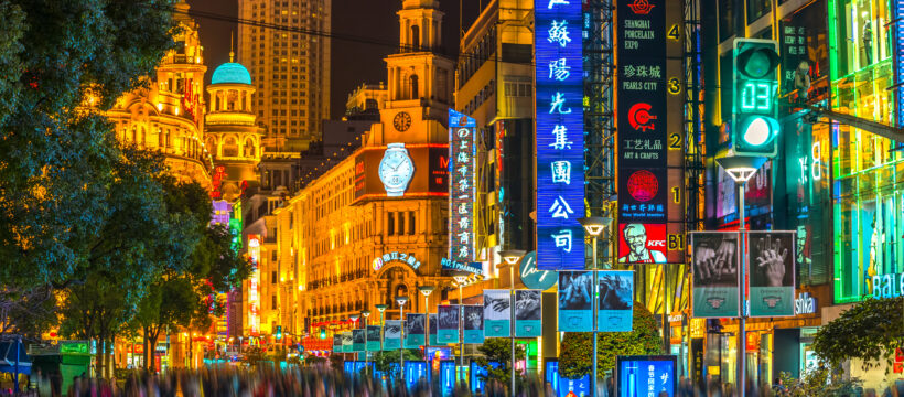 Busy neon-lit city street at night with tall colorful signs and blurred pedestrians getting by the storefronts.