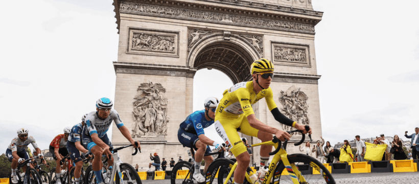 Group of pro cyclists racing past the Arc de Triomphe in Paris, yellow-clad rider leading on a cobblestone street