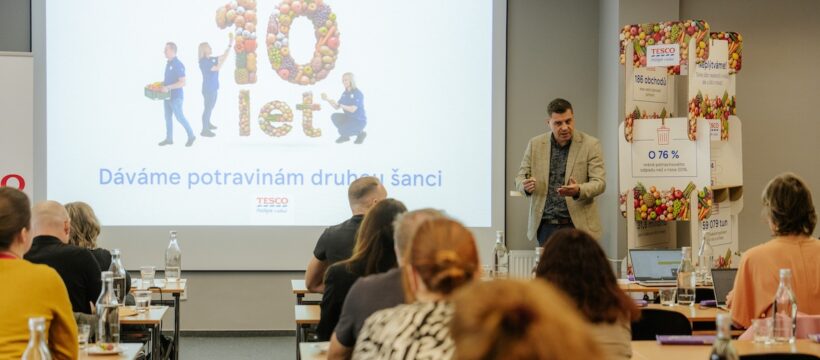 Speaker presenting a Tesco event in a conference room, large screen shows